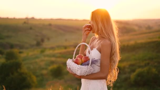 Krásná sexy blondýnka v bílých šatech na poli při západu slunce s košíkem s ovocem 