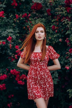 Kısa kırmızı elbiseli güzel kızıl saçlı kız. Yaz mevsiminde çiçeklerle bahçede poz vermek.