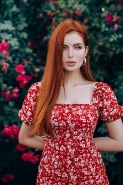 Kısa kırmızı elbiseli güzel kızıl saçlı kız. Yaz mevsiminde çiçeklerle bahçede poz vermek.