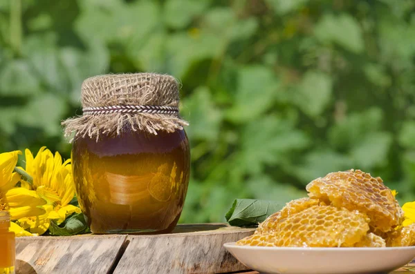 Frascos de miel, panal de abeja sobre mesa de madera con flores de fondo — Foto de Stock