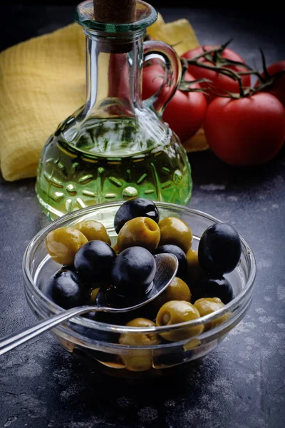 Green and black olives in bowl on black surface in front of oil bottle.