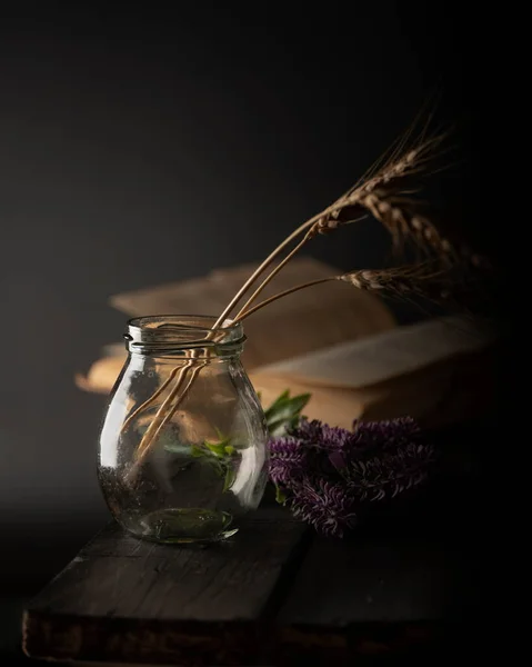Dark moody still life with book, flower and glass bottle with ears of wheat on wood table. Stok Gambar