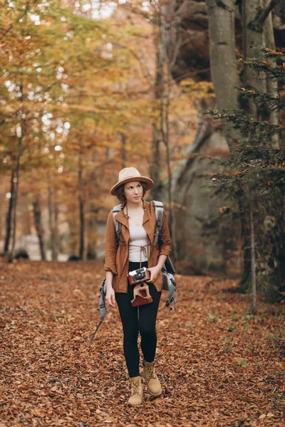 Attraktive junge Frau mit stylischem Hut und Reisetasche auf den Schultern, die sich im reizvollen Herbstwald umsieht — Stockfoto
