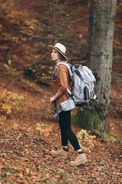 Young woman in a stylish hat and travel bag on her shoulders, looking around at the charming autumn forest — Stock Photo, Image