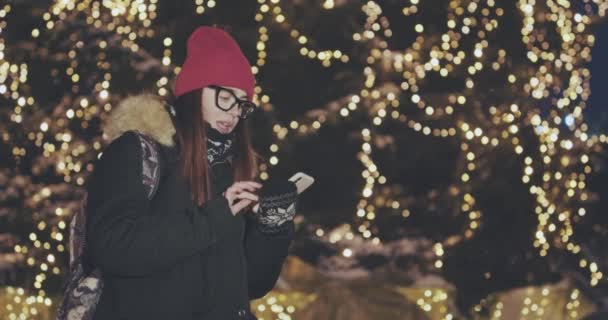 年轻美丽的女人用智能手机手举行户外在城市的夜晚, 微笑着, 脸上照亮了屏幕灯 — 图库视频影像