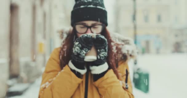 Дівчина в окулярах гуляє по місту і п'є гарячу каву. Усміхнена жінка п'є молочний коктейль під час прогулянки по зимовій міській вулиці. Портрет красивої брюнетки крупним планом — стокове відео