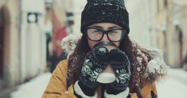 Hermosa chica caminando por la ciudad de invierno con una taza de café, mirando a la cámara y sonriendo — Vídeos de Stock