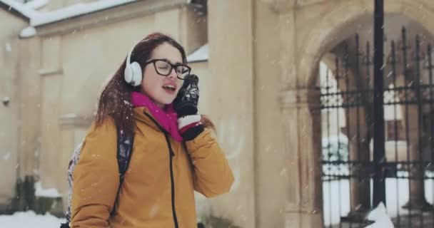 Bastante atractiva chica caucásica perforada en la ciudad nevada. Está escuchando música con grandes auriculares blancos. Caminatas al aire libre — Vídeos de Stock