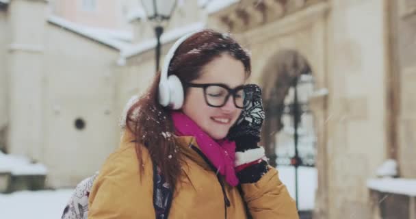 Retrato de cerca de una linda niña sonriente saliendo del clima nevado, escuchando música con sus auriculares inalámbricos — Vídeo de stock