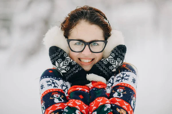 Portrait of young beautiful girl wearing ear muffs, sweater posing in snowy park. Woman looking camera and smiling — Stock Photo, Image