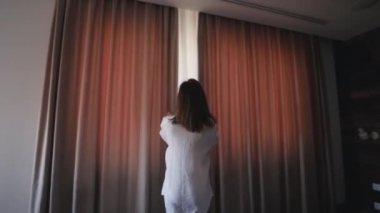 İnce, genç bir kadın büyük pencerenin yanında duruyor, ellerini uzatıyor, sabah güneş doğarken uyanıyor. Kadın, uyan ve sabah temiz hava almak için perdeleri aç.