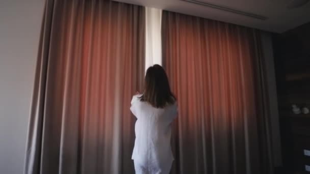 Jovem mulher esbelta fica perto da grande janela, estende as mãos para cima, acordar de manhã durante o nascer do sol. Mulher, acorda e abre as cortinas de manhã para apanhar ar fresco. — Vídeo de Stock