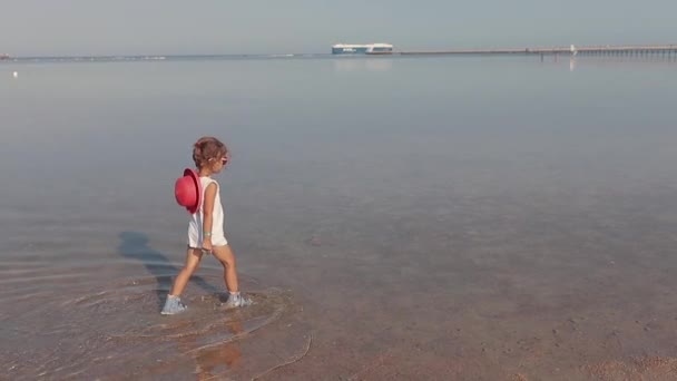 Aranyos kislány fehér ruhában megy el a homokos tengerpart mentén lassítva — Stock videók