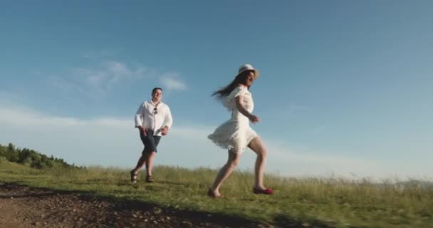 Alegre joven pareja de senderismo en verano en la cima de la montaña. Amor en las montañas. Hombre y mujer corren a lo largo del camino hacia el horizonte en algún lugar de la colina mientras el sol brilla sobre ellos — Vídeo de stock