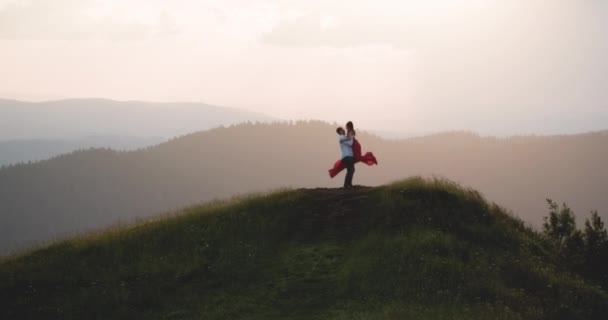 恋に落ちた若いカップルが山の上で夕日を楽しんでいます。緑の密集した山の地域でお互いにキスし、抱きしめる若い愛らしいカップルの素晴らしいlビュー。夏、夕焼け。永遠の恋に — ストック動画