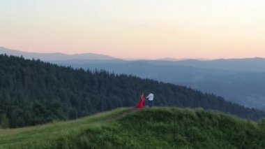 Aşık genç çift dağların üzerinde gün batımının tadını çıkarıyor. Kırmızı elbiseli kız. Genç sevimli çift inanılmaz havadan görünümü kucaklayan, dağlarda yeşil yoğun birbirlerini öpme
