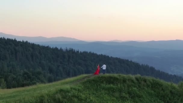 Aşık genç çift dağların üzerinde gün batımının tadını çıkarıyor. Kırmızı elbiseli kız. Genç sevimli çift inanılmaz havadan görünümü kucaklayan, dağlarda yeşil yoğun birbirlerini öpme — Stok video