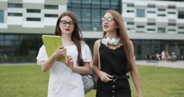 İki beyaz güzel genç kız öğrenci yürüyor ve bir kız diğerine bazı kitapları gösteriyor ve onları nereye götürdü. Portre çekimi. Okulda yürüyen mutlu öğrenciler.