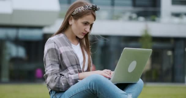 Close-up retrato de uma menina inteligente trabalhando em seu laptop ao ar livre enquanto sentado no gramado contra o grande edifício — Vídeo de Stock