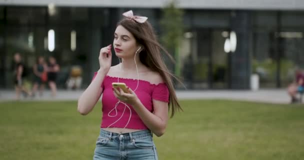 Hermosa joven usando un teléfono inteligente, escribiendo mensajes, escuchando música, caminando en el parque — Vídeos de Stock