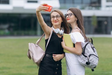 Yazlık kıyafetler giyen iki genç gülümseyen hipster kadın. Akıllı telefonda selfie selfie portre fotoğrafları çeken kızlar. Sokak arka planında poz veren mankenler. Kadın olumlu yüz duyguları gösteren