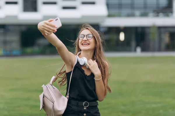 Munter ung studerende pige transporterer rygsæk udendørs, tager selfie. Billede af glad ung smuk kvinde studerende gå i parken tage en selfie ved kamera - Stock-foto