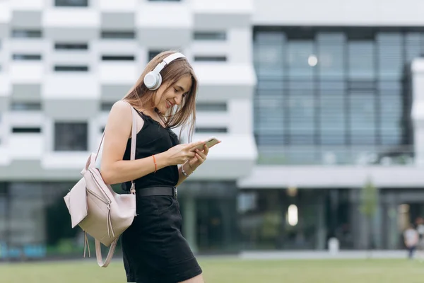 Attraktiv pige lytter musik fra telefonen i hovedtelefoner, skrive beskeder, mens du går på den urbane gade. Livsstil . - Stock-foto