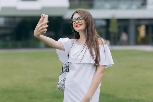 휴대 전화 통화를 사용하여 자연 공원에서 벤치 야외에서 안경을 쓰고있는 학생 소녀가 셀카를 찍습니다.. — 스톡 사진