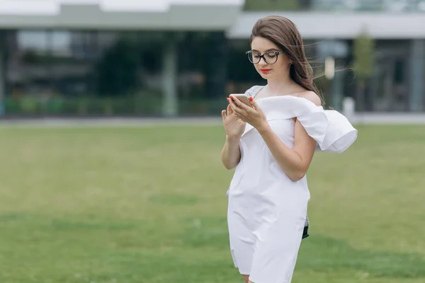 Feliz mujer de negocios usando smartphone con camisa azul y gafas. Joven empresaria atractiva alegre en gafas de pie y usando el teléfono móvil —  Fotos de Stock