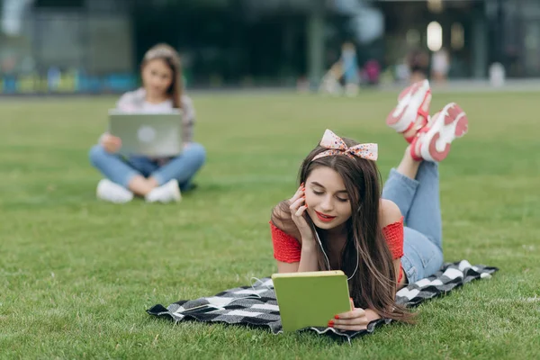 Dziewczyna czytając książkę w parku, siedząc na trawie i odpoczywać w kampusie uniwersyteckim. Kobieta słuchanie muzyki przez zestaw słuchawkowy i czytanie książki w parku — Zdjęcie stockowe