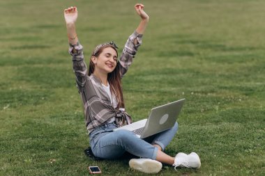 Güzel rahat kadın dizüstü bilgisayar kullanarak çim üzerinde oturan. Parkta yeşil çimenler üzerinde oturan çekici genç kadının portresi