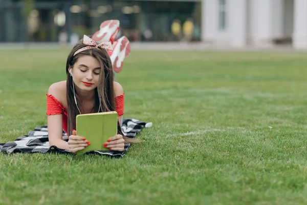 Uśmiechnięta młoda kobieta leżąca na zielonej łące i słuchając muzyki przez słuchawki. Kobieta słucha muzyki — Zdjęcie stockowe