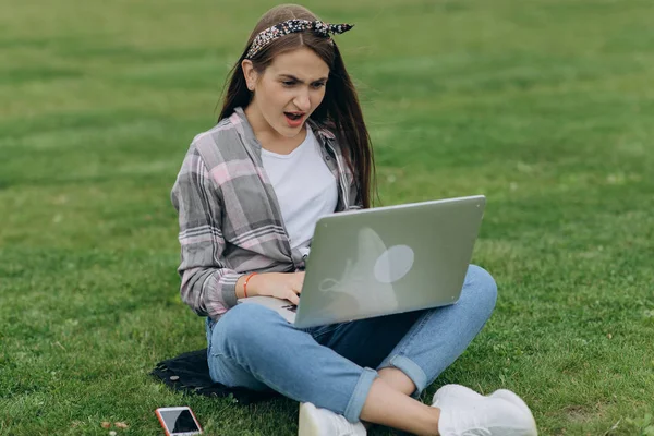 Sociable young woman sitting on green grass in park with legs crossed during summer day while using laptop for video call — Stock Photo, Image