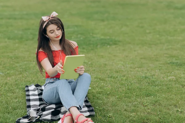 Kobieta słuchanie muzyki przez zestaw słuchawkowy i czytanie książki w parku — Zdjęcie stockowe