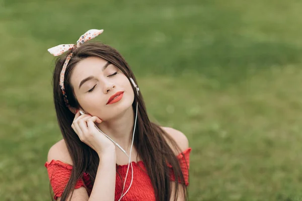 Primo piano ritratto di contenuto giovane bella donna con le cuffie, ascoltare musica con gli occhi chiusi e sdraiata sull'erba nel parco — Foto Stock