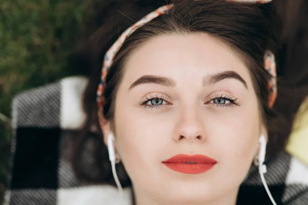 Retrato de cerca del contenido de la joven hermosa mujer con auriculares, escuchando música con los ojos abiertos y acostada en el césped en el parque. Vista superior . —  Fotos de Stock