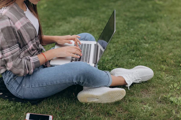 Attractive young woman in plaid shirt sitting on green grass in park during summer day while using laptop — Stock Photo, Image