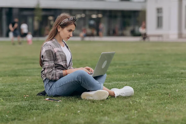 Portrait of young woman sitting in park with legs crossed during summer day while using laptop — Stock Photo, Image