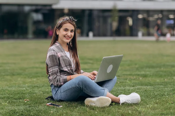 Pretty young woman sitting on green grass in park with legs crossed during summer day while using laptop — Stock Photo, Image