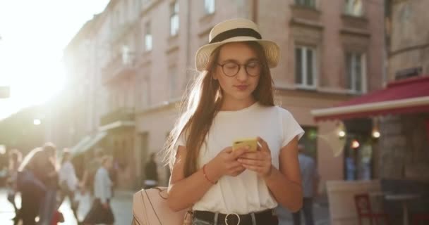 Atraktivní dívka žvanila na jejím mobilním zařízení. Rozhlížel se kolem. Chodící do města. Nosí slaměný klobouk. Má elegantní účes. Žena s krásnou make-up — Stock video