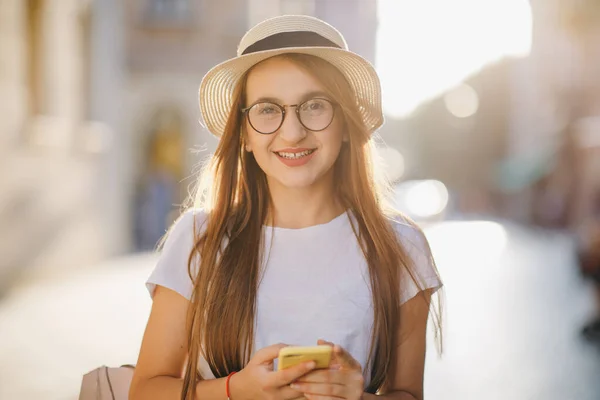 Jeune femme voyage en chapeau et lunettes se tient sur la rue de la ville et SMS SMS sur son téléphone mobile. Attrayant jeune femme dans un look élégant en utilisant APP sur son iPhone. Réseaux sociaux. Être en ligne . — Photo