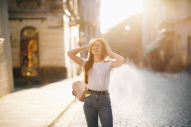 Sırt çantası ve gözlükleriyle şapkalı mutlu ve seksi bir kadının portresi. Gün batımını seyrediyor. Gülümsüyor ve şehirde dolaşıyordu. Seyahat ve yaşam tarzı. Yaz zamanı. Pozitif
