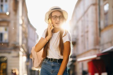 Güneşli bir sonbahar günü, arka plan. Genç ve çekici bir kadın şapka ve gözlüklerle şehir caddesinde duruyor, cep telefonuyla konuşuyor, gülümsüyor, gülüyor. Hippi kız yürüyor. Tatil, macera, seyahat