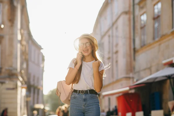 Módní hipster dívka ve stylovém klobouku, mluví s přítelem přes mobil, sdílí šťastné emoce po objevování nových památek ve městě, stojí nad budováním venkovní — Stock fotografie