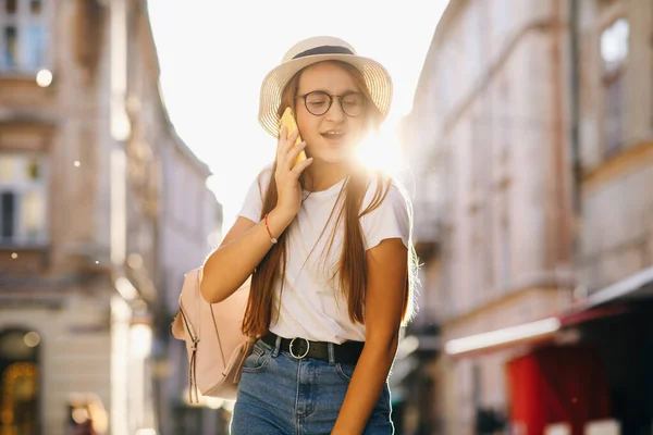 Ηλιόλουστη φθινοπωρινή μέρα, πίσω φως. Νεαρή ελκυστική γυναίκα ταξιδεύει με καπέλο και γυαλιά στέκεται στην οδό Σίτι, μιλώντας στο κινητό, χαμογελώντας, γελώντας. Hipster κορίτσι περπατά. Διακοπές, περιπέτεια, ταξίδι — Φωτογραφία Αρχείου