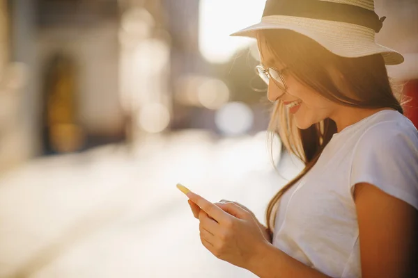 Heureuse belle femme marchant et écrivant ou lisant des messages SMS en ligne sur un téléphone intelligent. Fille en chapeau et lunettes. SMS de messages . — Photo