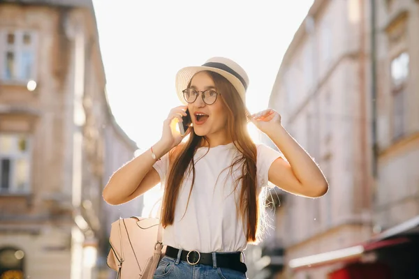 Woman in hat and eyeglasses stands on city street, talking on cell phone, smiling, laughing. Hipster girl walks. Vacation, adventure, trip. Sunny day, backlight.