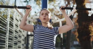 Genç kız sonbahar parkında barfiks egzersizi yapıyor. Spor kıyafetli çekici genç kaslı kız barfiks egzersizi yapıyor ve başını dik tutuyor. Genç Kadın Spor Malzemeleriyle Egzersiz Yapıyor