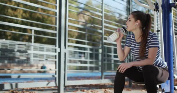 Sportgetränk, junge Frau trinkt Mineralwasser nach Ausdauertraining in der Natur bei strahlendem Sonnenschein. Mädchen sitzt müde nach dem Training auf der Bank. Trinken aus dem Shaker. — Stockvideo