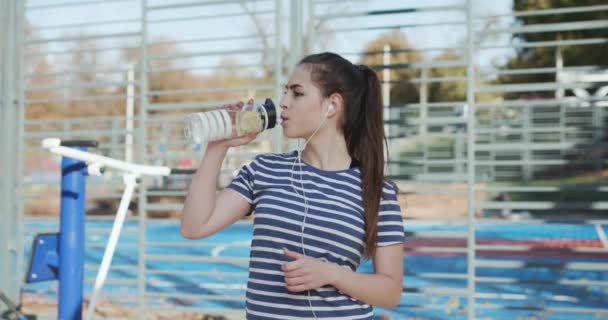 A menina está cansada depois de treinar bebidas de uma garrafa. Mulher bonita está bebendo uma bebida de uma coqueteleira no fundo de equipamentos esportivos . — Vídeo de Stock